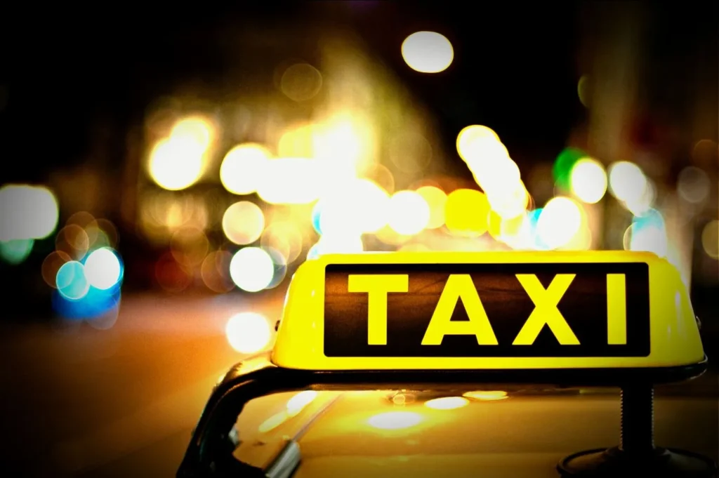 A close-up of a yellow taxi sign illuminated at night, mounted on top of a vehicle from a reputable taxi service in Trivandrum. The background is blurred with colorful, out-of-focus lights, creating a vibrant, bokeh effect.